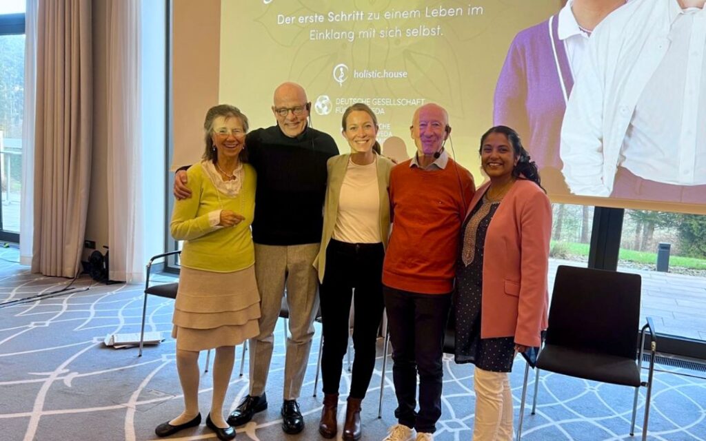 Dozierende der Ayurveda Fach-Coach Ausbildung stehen Arm in Arm nebeneinander und lächeln in die Kamera. Im Hintergrund die Titelfolie eines Vortrags.