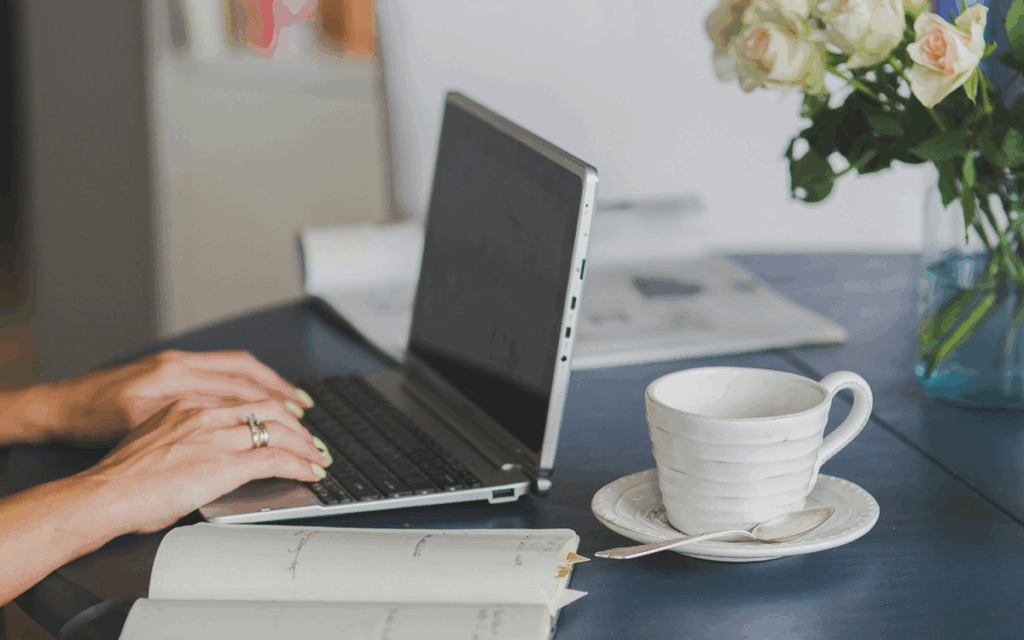 Hände einer Frau an einem Laptop daneben ein aufgeschlagenes Notizbuch und eine Tasse Tee